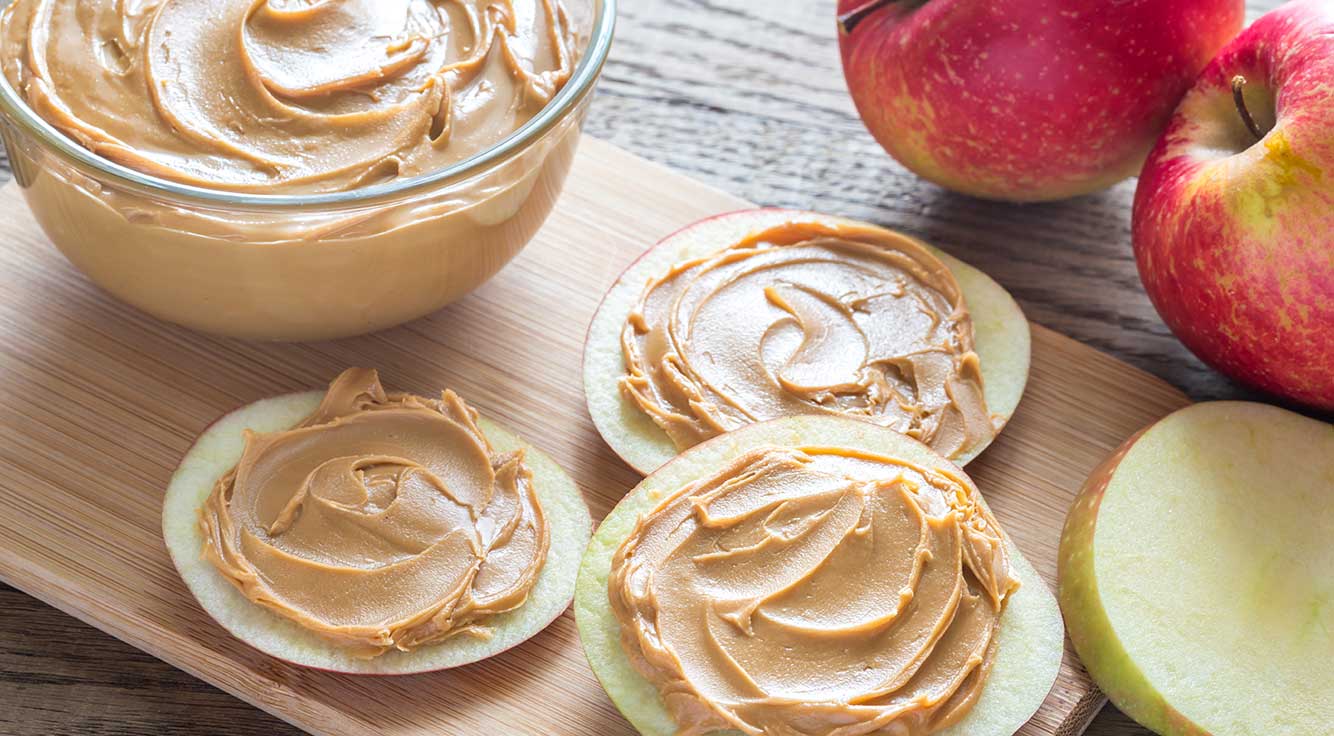 Cut apple slices topped with smooth peanut butter on a cutting board by a bowl of peanut butter and 2 red apples.