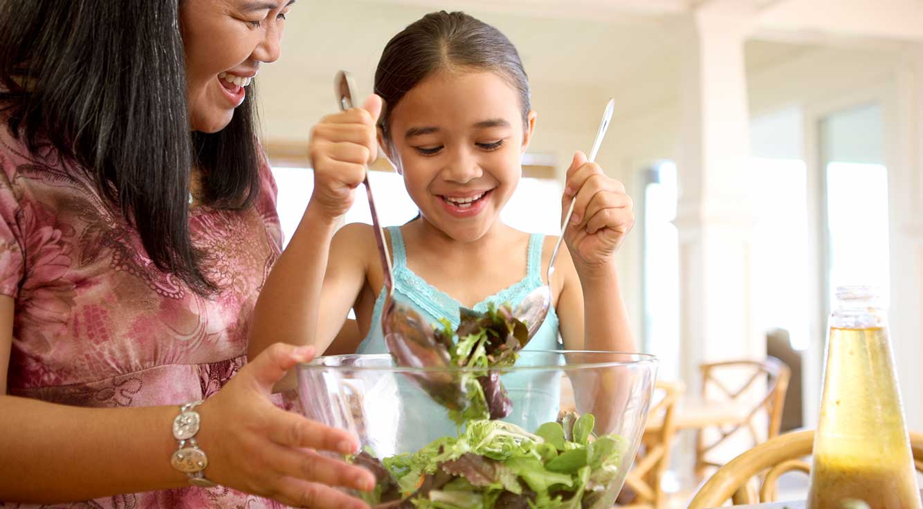 A smiling mom watches her young daughter use tongs to grab some salad from a bowl.