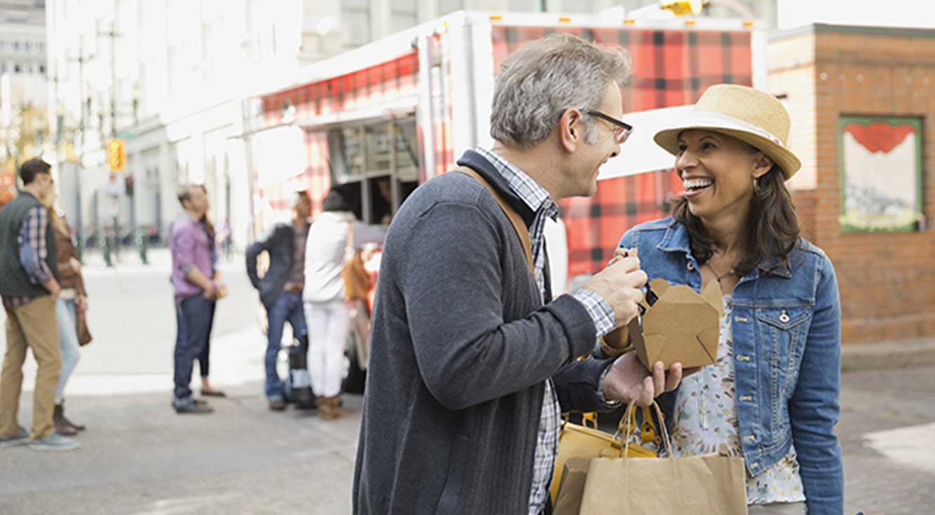 A smiling middle-age couple share a meal from a food truck on a city street
