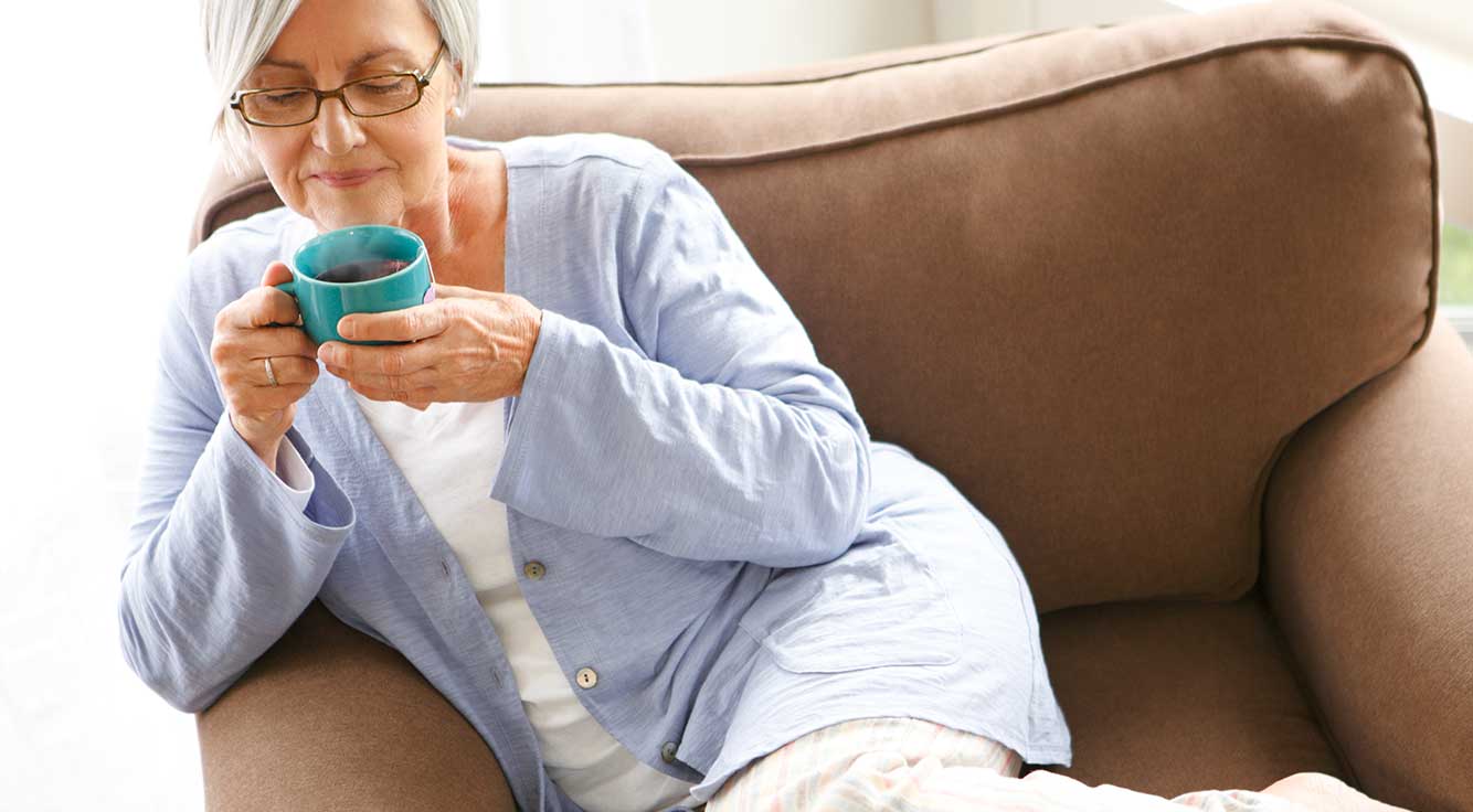 An older woman enjoys a cup of tea on the couch.