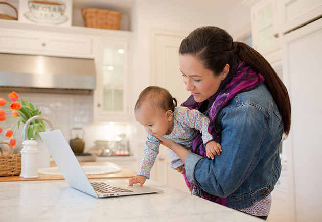 Woman looking at laptop while holding young child at home.